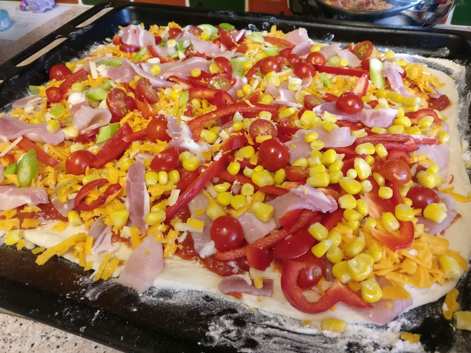 A tray pizza topped with bacon, sweetcorn, peppers, cherry tomatoes and grated cheese before baking