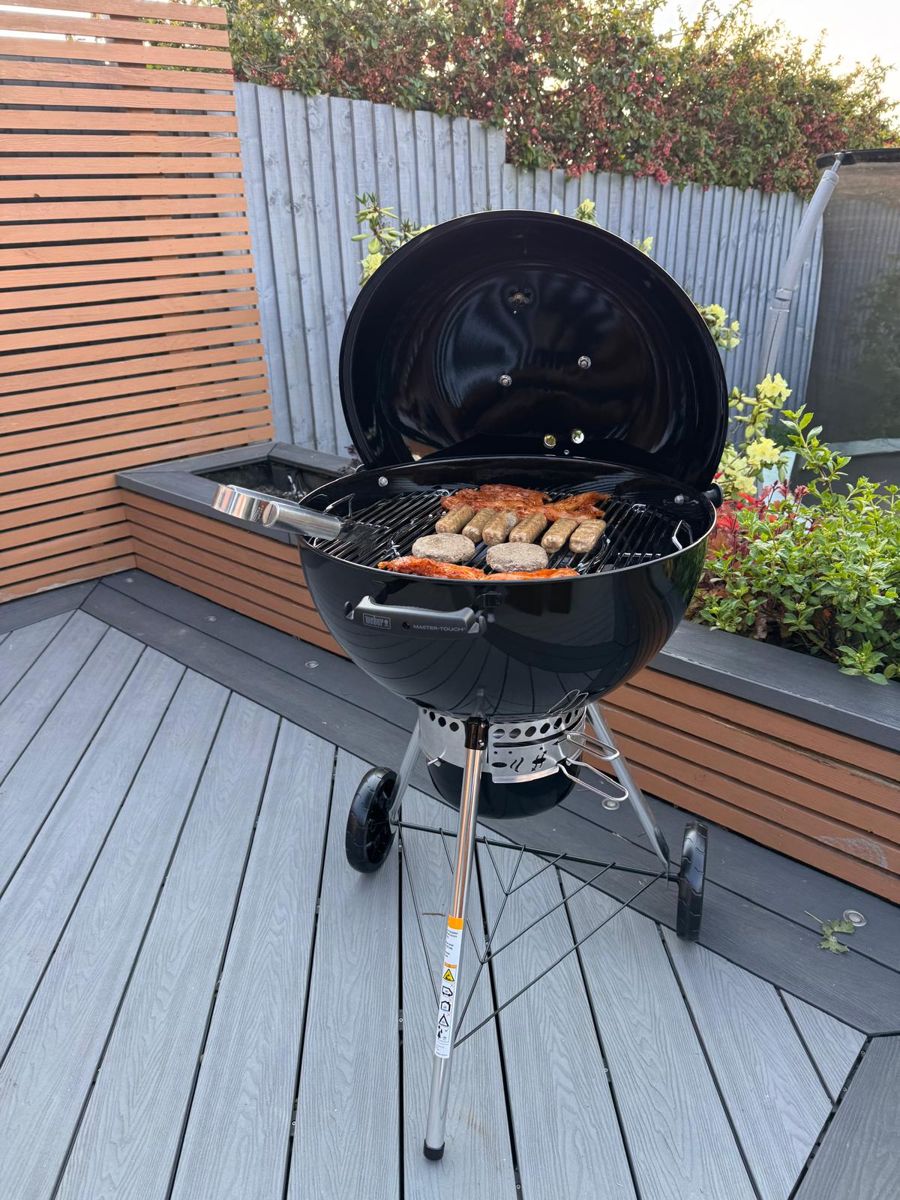 Weber Master-Touch kettle BBQ loaded with food on a grey composite deck with slatted fencing